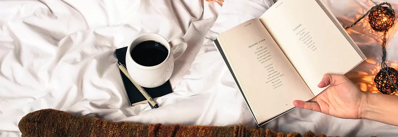A mug of tea, a book and a pair of legs stretched out on a bed.