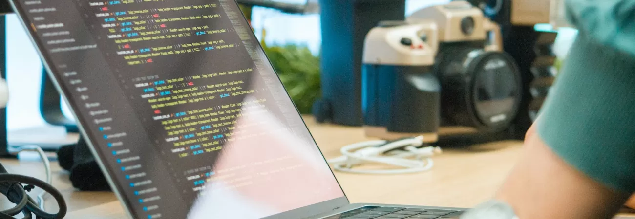 A person leaning over a desk and a laptop with computer code on the screen.